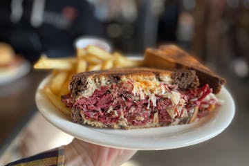 A hand holds a plate with a Rueben: marble rye bread, thick layers of corned beef with visible sauerkraut and thousand island dressing in the Urban Gr